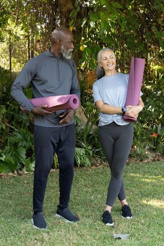 Diverse mature couple carrying purple yoga mats in backyard garden practicing wellness