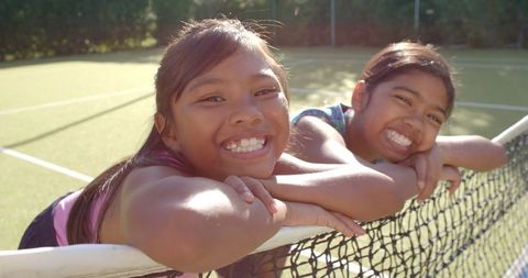 Smiling girls leaning on tennis net embracing youthful joy
