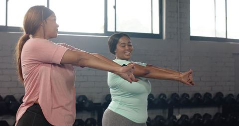 Two diverse women stretching arms together smiling in daylight gym wearing leggings