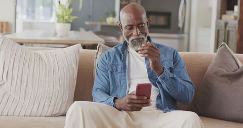 Senior man sitting on sofa reacting to smartphone notification while tapping red phone