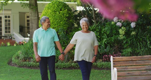 Senior couple enjoying a walk in a lush garden, holding hands, and smiling at each other. Ideal for use in advertisements, brochures, or websites promoting senior lifestyle, health, retirement communities, or romantic relationships in later life.