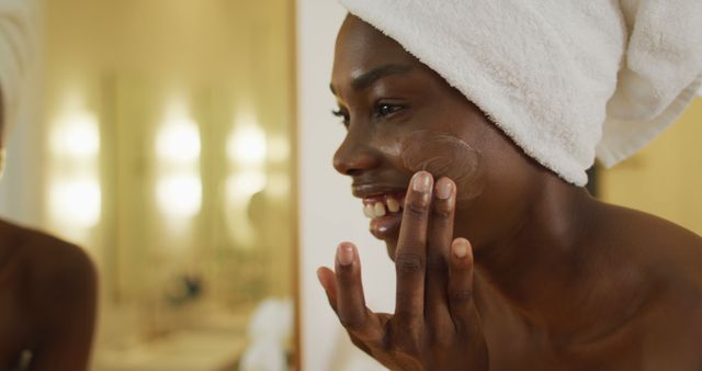 This image features a smiling woman with a towel wrapped around her head applying face cream in a bathroom. It showcases a skincare routine, emphasizing beauty, self-care, and wellness. Ideal for use in articles, advertisements, or marketing materials related to beauty products, skincare routines, personal grooming, and lifestyle wellness tips.