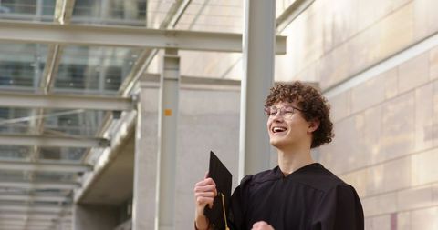 Young graduate celebrating with mortarboard by modern glass canopy, urban architecture
