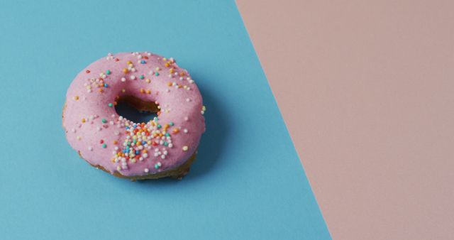 Close-up of a single pink frosted donut with colorful sprinkles on half blue, half pink pastel background. Perfect for illustrating bakery products, dessert menus, or social media content related to sweets and treats.