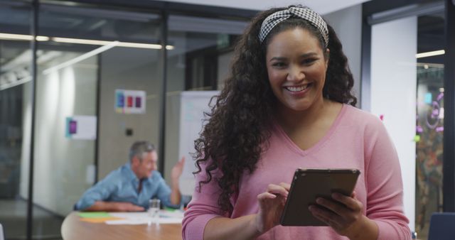 Businesswoman with curly hair smiling while using tablet in modern office, with a colleague in the background. Perfect for promoting workplace technology, professional settings, teamwork, and modern business environments.