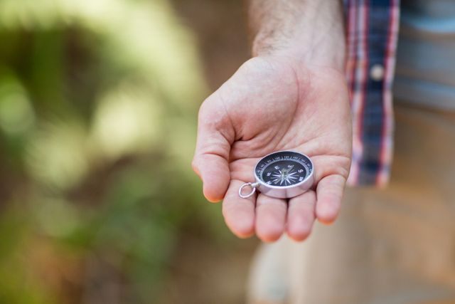 Close-up of a hiker's hand holding a compass in the countryside, ideal for themes related to navigation, outdoor adventure, and exploration. Useful for travel blogs, hiking guides, camping equipment advertisements, and nature exploration articles.