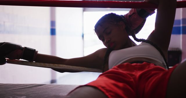 Female boxer in bright athletic wear resting with boxing gloves on after intense training in gym. Perfect for content about sports training, fitness determination, women in sports, boxing equipment promotion, or motivational themes.