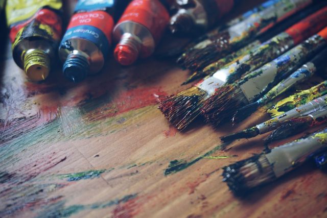 Messy paintbrushes and paint tubes are scattered on a wooden surface, creating a colorful and creative scene. Ideal for depicting art, creativity, and artistic processes. Perfect for illustrating articles or blogs related to painting, art supplies, art studios, and creative activities.