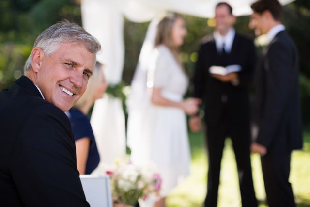 Man smiling while attending an outdoor wedding ceremony in a park. Ideal for use in wedding invitations, event planning brochures, or articles about wedding celebrations and guest experiences.