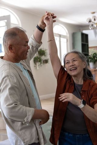 Senior asian couple dancing joyfully twirling and smiling in sunlit living room