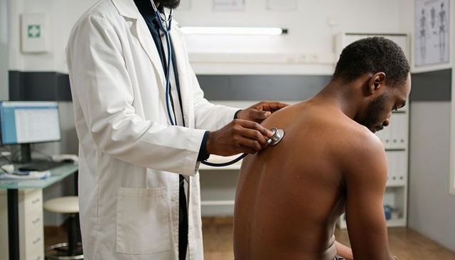 Physician listening to patient lungs with stethoscope on bare back in clinic exam room