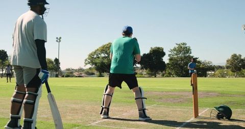 Batsman standing at crease wearing pads and helmet on sunny grass cricket pitch with fielders