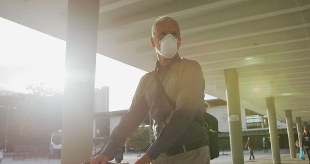 Older man wearing protective face mask using bicycle in urban area during sunset. Ideal for content related to health and safety, commuting, active lifestyle of senior citizens, and urban living. Bright sunlight in the background adds a hopeful tone.