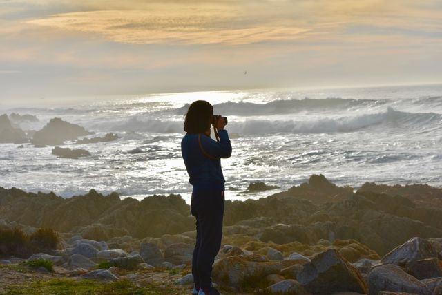 Woman holding camera and taking photos of ocean waves with sun setting on horizon. Ideal for travel magazines, coastal landscape promotions, and hobby photography blogs.