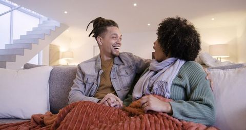 African american couple holding hands on sofa under cozy blanket smiling and laughing