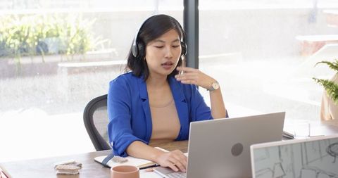 Professional Asian Woman in Office Wearing Headset and Working on Laptop