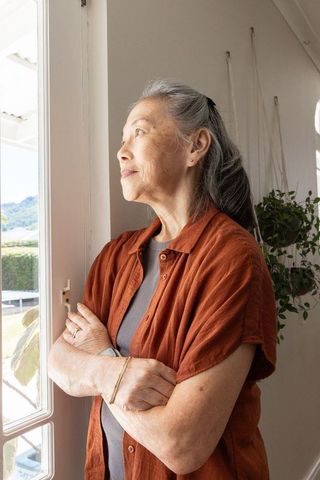 Senior asian woman standing by glass door looking outside, calm contemplative home moment
