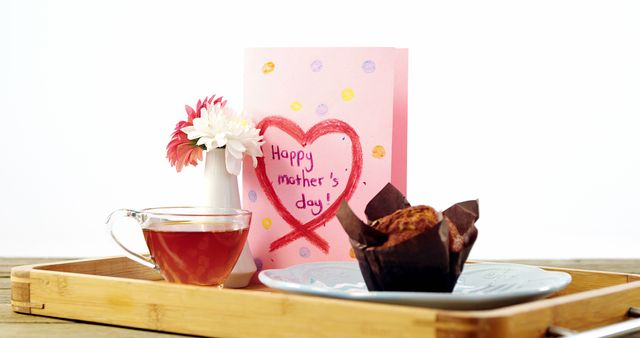 Photo features a Mother’s Day card with a pink heart design, placed on a wooden tray alongside a clear glass of tea, a decorative pink flower, and a muffin in wrapping paper. Perfect for use in advertisements, blogs, or social media posts celebrating Mother’s Day.