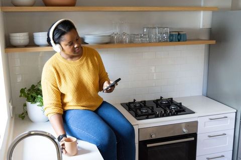 African american woman sitting on kitchen counter wearing headphones using smartphone