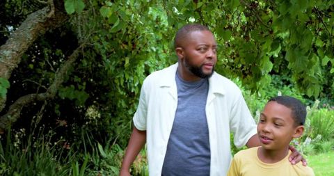 African american father guiding son through lush green backyard under tree canopy