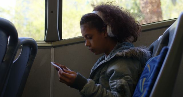 Young girl sitting on a bus, wearing headphones and looking at a handheld device. Perfect for depicting themes of public transportation, youth using technology, concentration, school commute, modern child lifestyle, and everyday urban life.