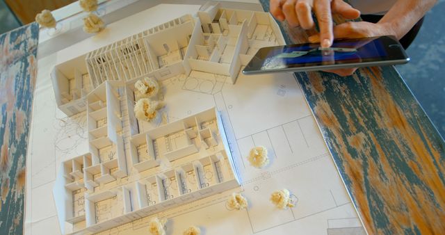 A top-down view of an architect consulting a blueprint and a 3D architectural model while interacting with a digital tablet. Great for illustrating themes related to architecture, planning, technology in construction, and professional consulting in design projects.