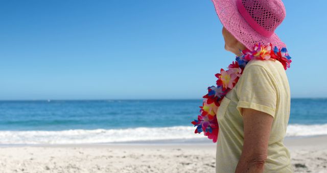 Senior woman enjoying sunny beach, wearing pink sun hat and colorful lei, facing ocean. Perfect for vacation, tropical, retirement lifestyle themes.