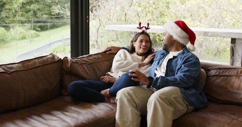 Couple relaxing on leather sofa wearing santa hat and reindeer antlers during holiday