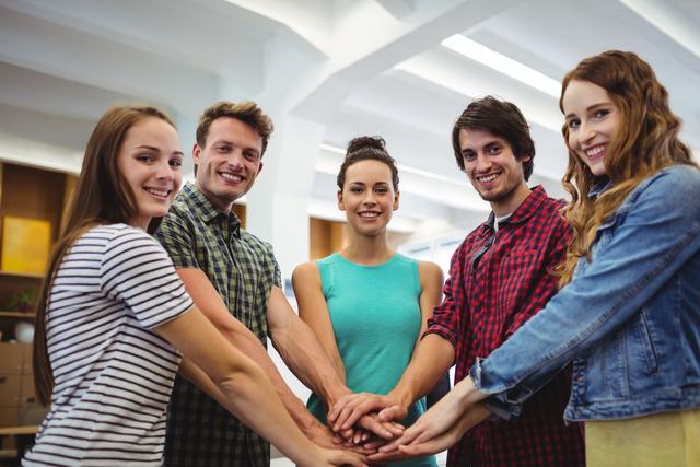 Group of young professionals standing in a modern office, stacking hands in a show of unity and teamwork. Ideal for illustrating concepts of collaboration, team spirit, and workplace motivation. Suitable for business presentations, team-building materials, and corporate websites.