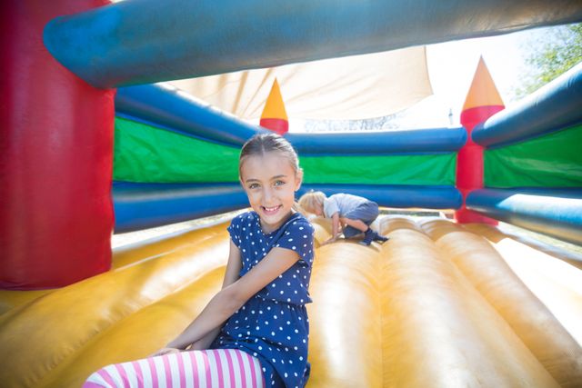 Happy girl sitting on a colorful bouncy castle while her brother plays in the background. Ideal for use in advertisements for family activities, playgrounds, summer events, and children's amusement parks. Perfect for illustrating joyful childhood moments and sibling fun.