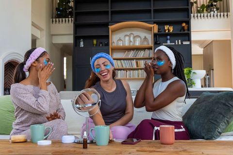 Friends sharing pamper night applying under-eye patches laughing on cozy living room sofa