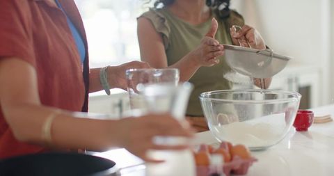 Diverse women sifting flour and measuring milk on bright home kitchen island