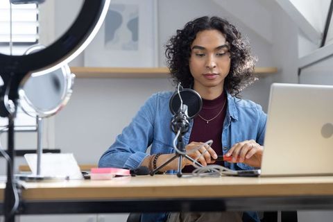 Non-binary creator adjusting microphone at laptop in home studio with ring light