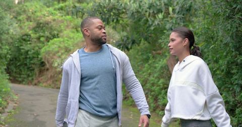 Diverse couple walking on park trail in hoodies showing wristwatch for active lifestyle