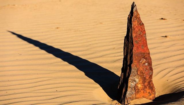Rust-streaked triangular monolith casting long shadow over rippled desert sand