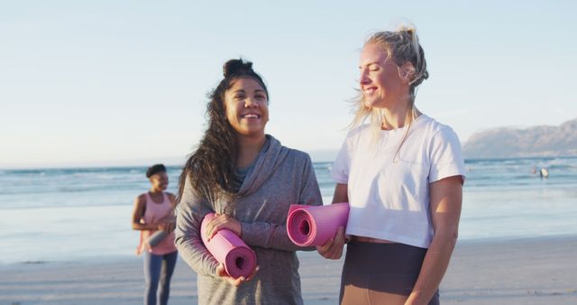 This stock photo shows two women smiling and holding yoga mats on a beach, with one person in the background by the ocean. It is ideal for promoting a healthy, active lifestyle, yoga classes, or fitness products. Perfect for use in wellness websites, social media campaigns, and fitness blog posts.