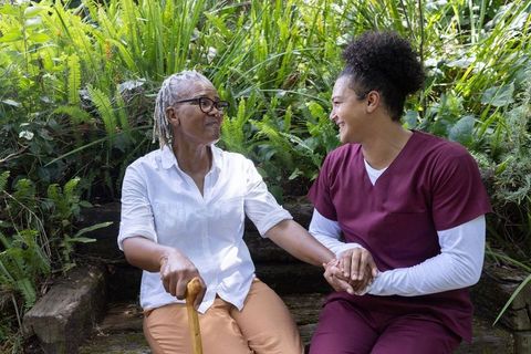 Elderly black woman holding cane, smiling and holding hands with caregiver on garden bench