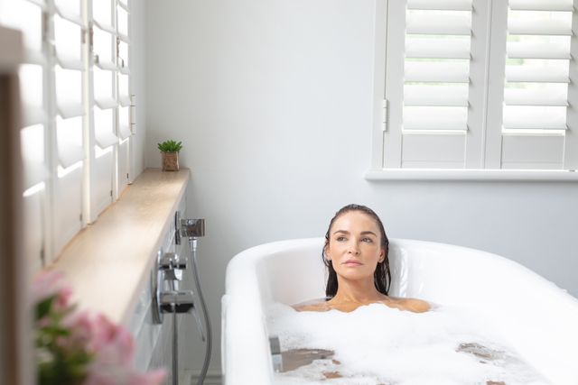 Woman enjoying bubble bath in white modern bathroom with soft natural lighting. Ideal for promoting self-care, relaxation, wellness, and home living concepts. Perfect for use in lifestyle blogs, home decor magazines, and wellness product advertisements.