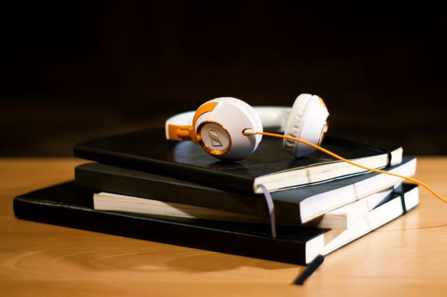 Close-up view of white and orange headphones resting on top of a pile of hardcover books on a wooden desk. The scene suggests a quiet study area or a library setting where one might combine studying with listening to music or educational content. Suitable for illustrating educational materials, study habits, or multimedia learning. Perfect for websites, blogs, or articles focused on education, learning, or study tips.