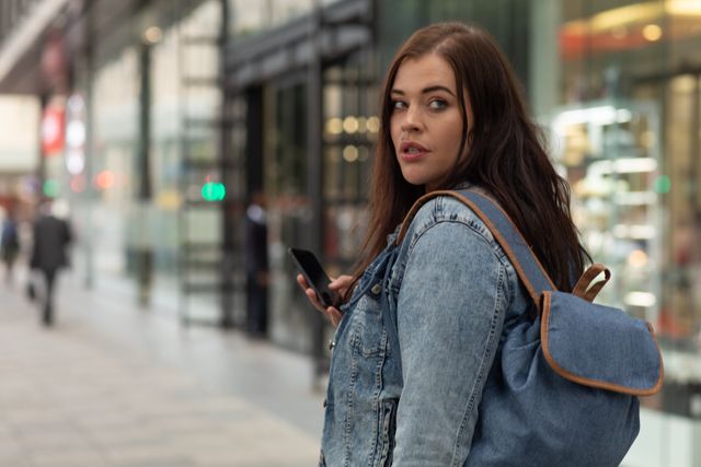Curvy Caucasian woman out and about in the city streets during the day, walking wearing a backpack, turning around while using her smartphone 