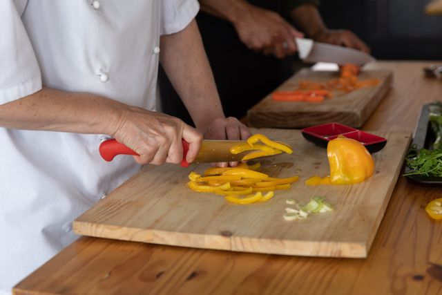 Caucasian female chef slicing yellow bell peppers on a wooden chopping board in a professional restaurant kitchen. Other chefs are working in the background, preparing various ingredients. Ideal for use in articles, blogs, or advertisements related to culinary arts, professional cooking, restaurant kitchens, and food preparation.