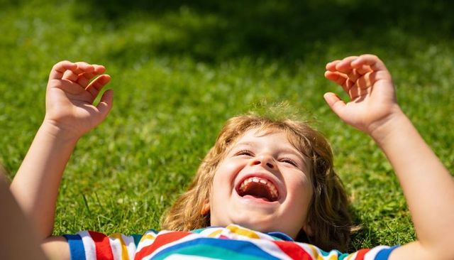 Laughing boy lying on green lawn wearing colorful striped t-shirt enjoying sunny summer joy