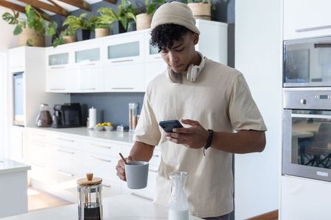 African american man making coffee and checking smartphone in modern minimalist kitchen