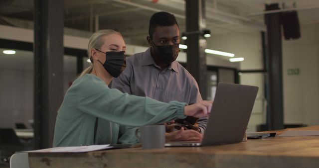 This scene depicts diverse coworkers, both wearing face masks, working together on a laptop in a modern office environment. The setting, featuring contemporary furnishings and safety measures, highlights the essence of teamwork in a professional setting during the Covid-19 pandemic. This visual can be used for contexts related to corporate teamwork, pandemic-era business operations, workplace diversity, and professional collaboration.