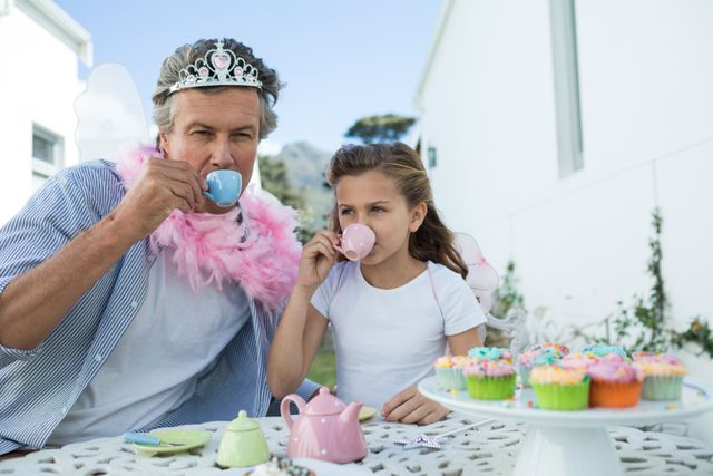 Father and daughter dressed in fairy costumes enjoying a tea party in a garden. The father wears a tiara and pink feather boa, while the daughter is sipping tea. Cupcakes and a tea set are on the table. Perfect for themes of family bonding, playful moments, childhood imagination, and outdoor activities.