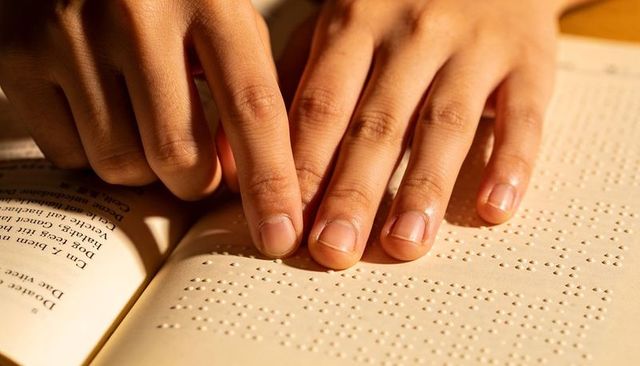 Adult hands reading braille with fingertips on embossed book page under warm golden light