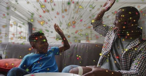 Black father and son celebrating high-five on couch with colorful confetti burst