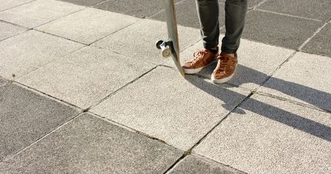 Skateboarder standing on tiled plaza in cuffed denim and brown sneakers casting shadow