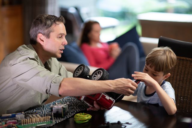Father and son working together to fix a remote controlled car at home, showcasing family bonding and teamwork. Mother is relaxing on a sofa in the background, adding a sense of a cozy family environment. Ideal for use in articles or advertisements about parenting, family activities, hobbies, and home life.