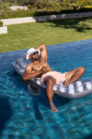 African american man relaxing on inflatable float with cocktail in sunny backyard pool